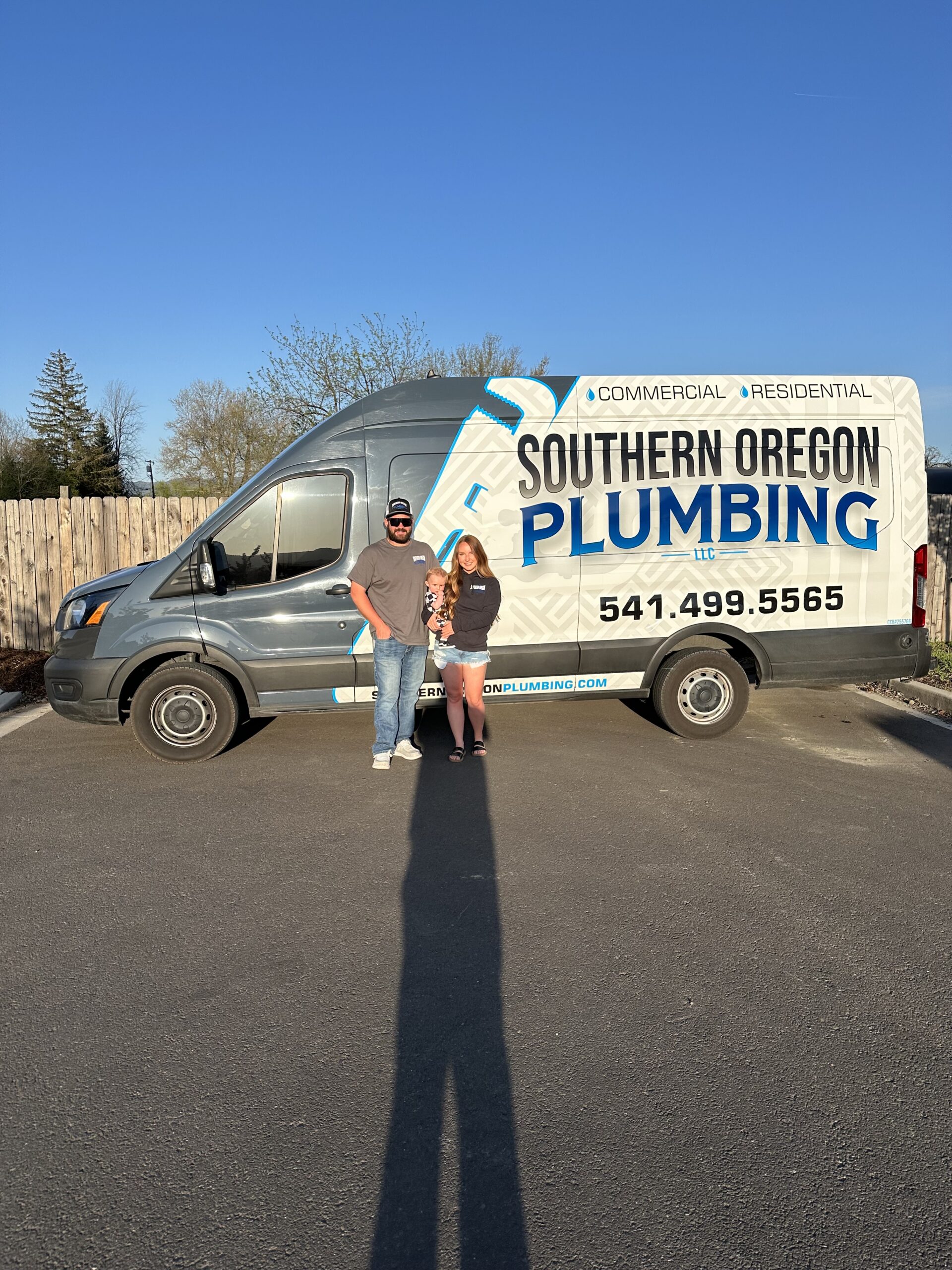 Two people stand in front of a Southern Oregon Plumbing van parked on pavement, under clear skies, with a wooden fence and trees in the background.