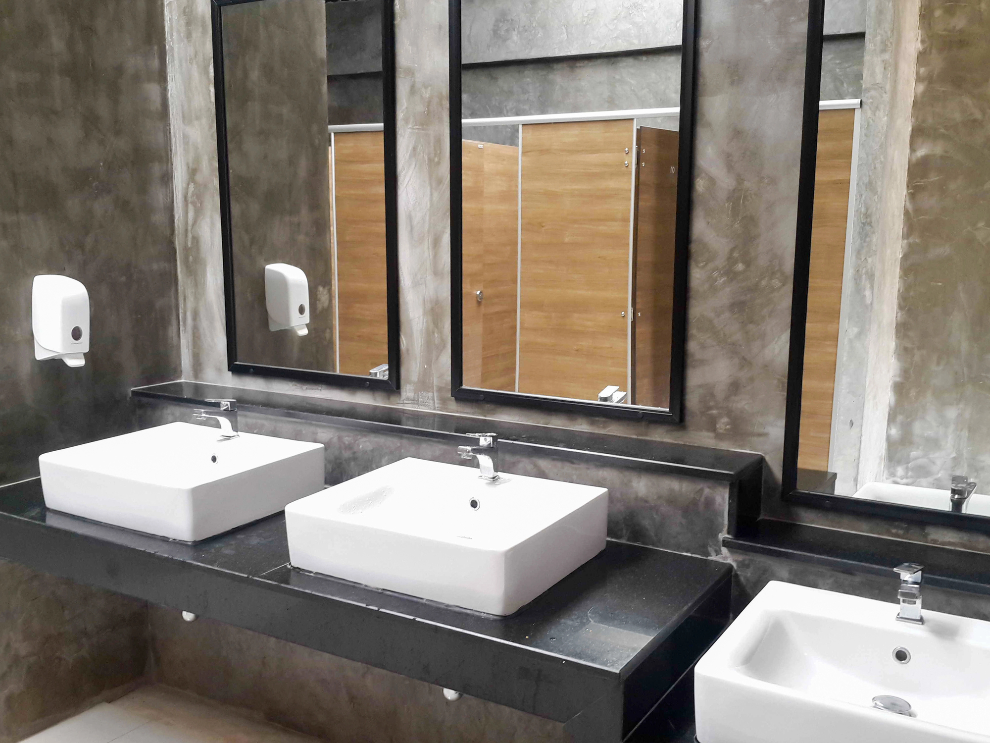 Modern public restroom with two white sinks, soap dispensers, large mirrors, and wooden stall doors in the background.