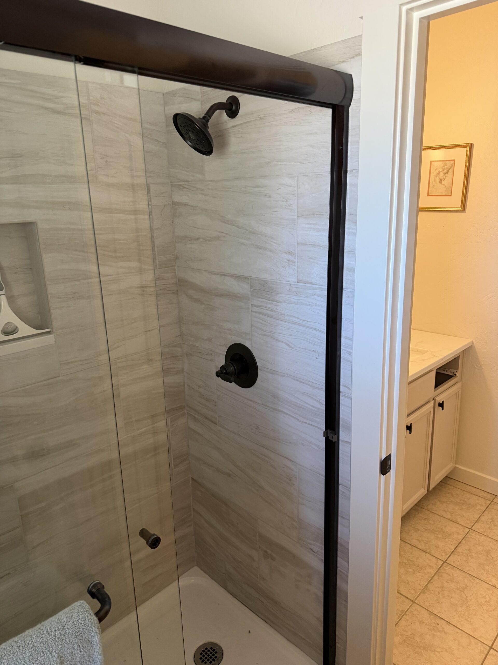 Shower with a glass door and dark fixtures next to a bathroom vanity area with cabinets and a towel hanging on the door.