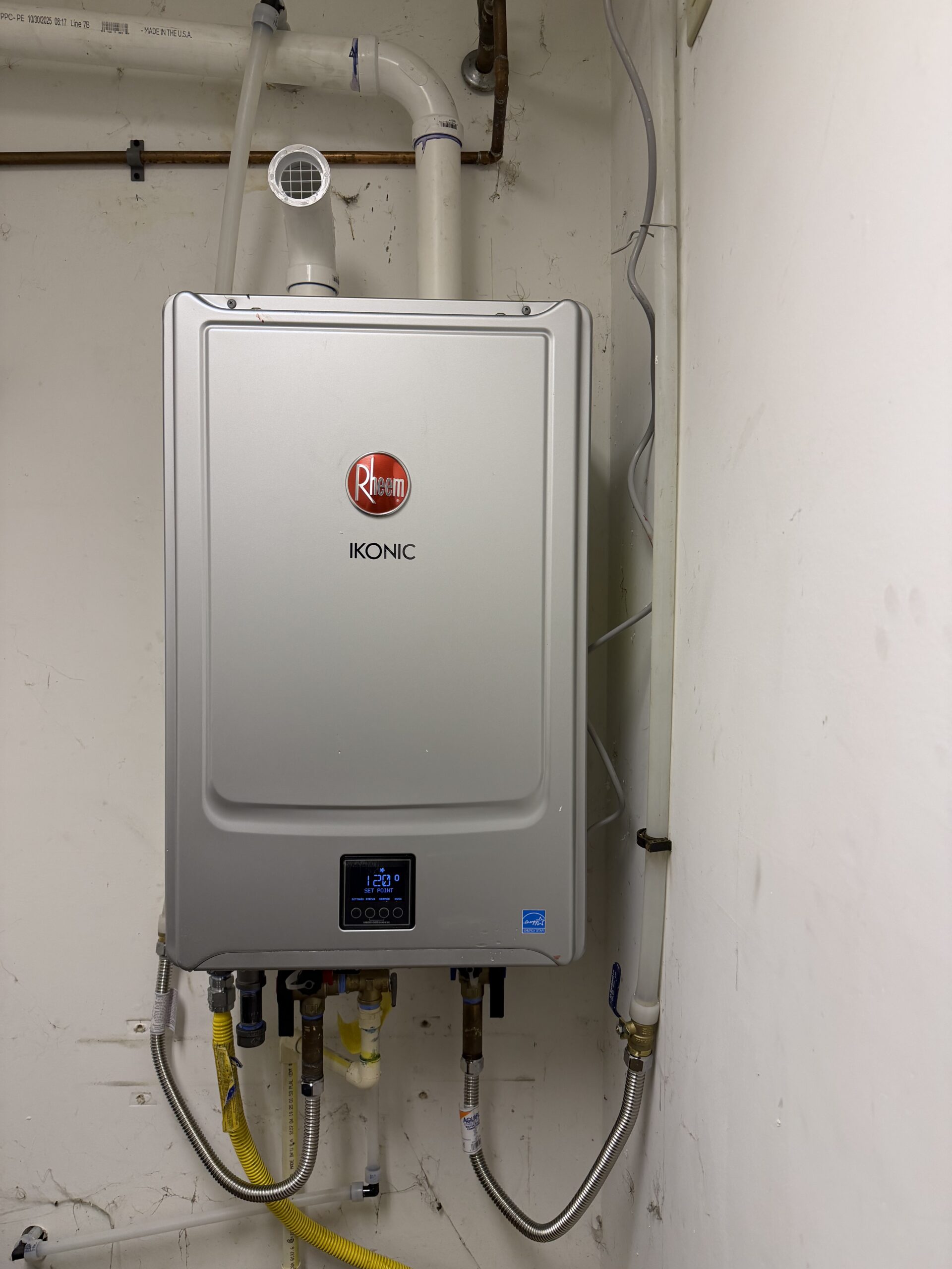 A Rheem Ikonic tankless water heater mounted on a wall, connected to various pipes and fittings in a utility room.