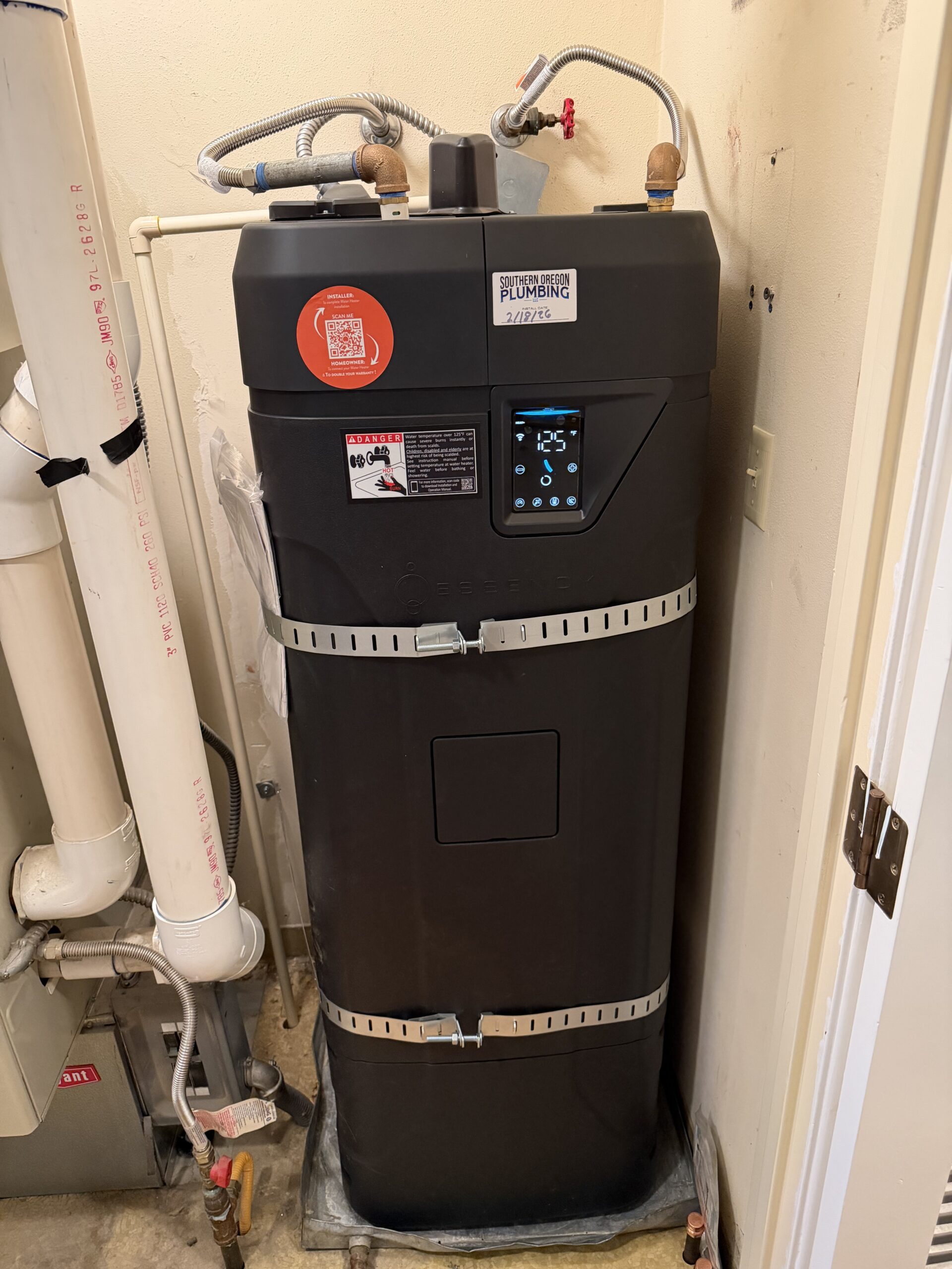 A black water heater displays a digital temperature reading of 125°F, with plumbing connections at the top, installed in a utility room next to white pipes.