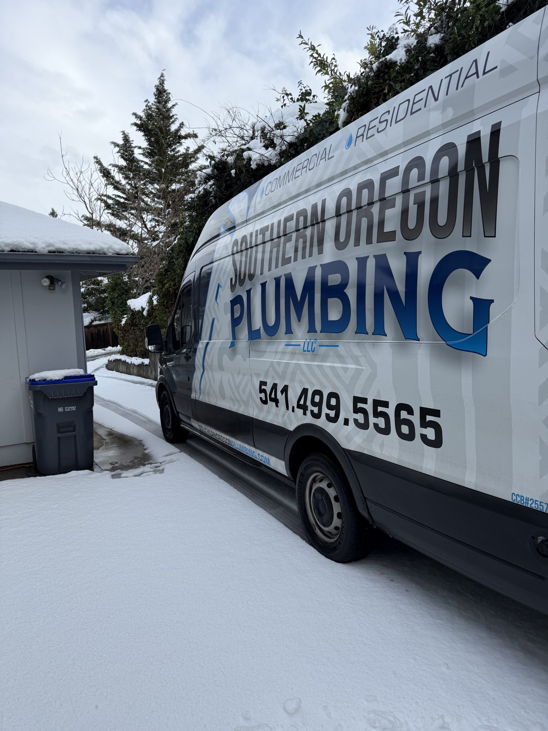 A Southern Oregon Plumbing van is parked on a snowy driveway beside a house with a trash bin near the garage.