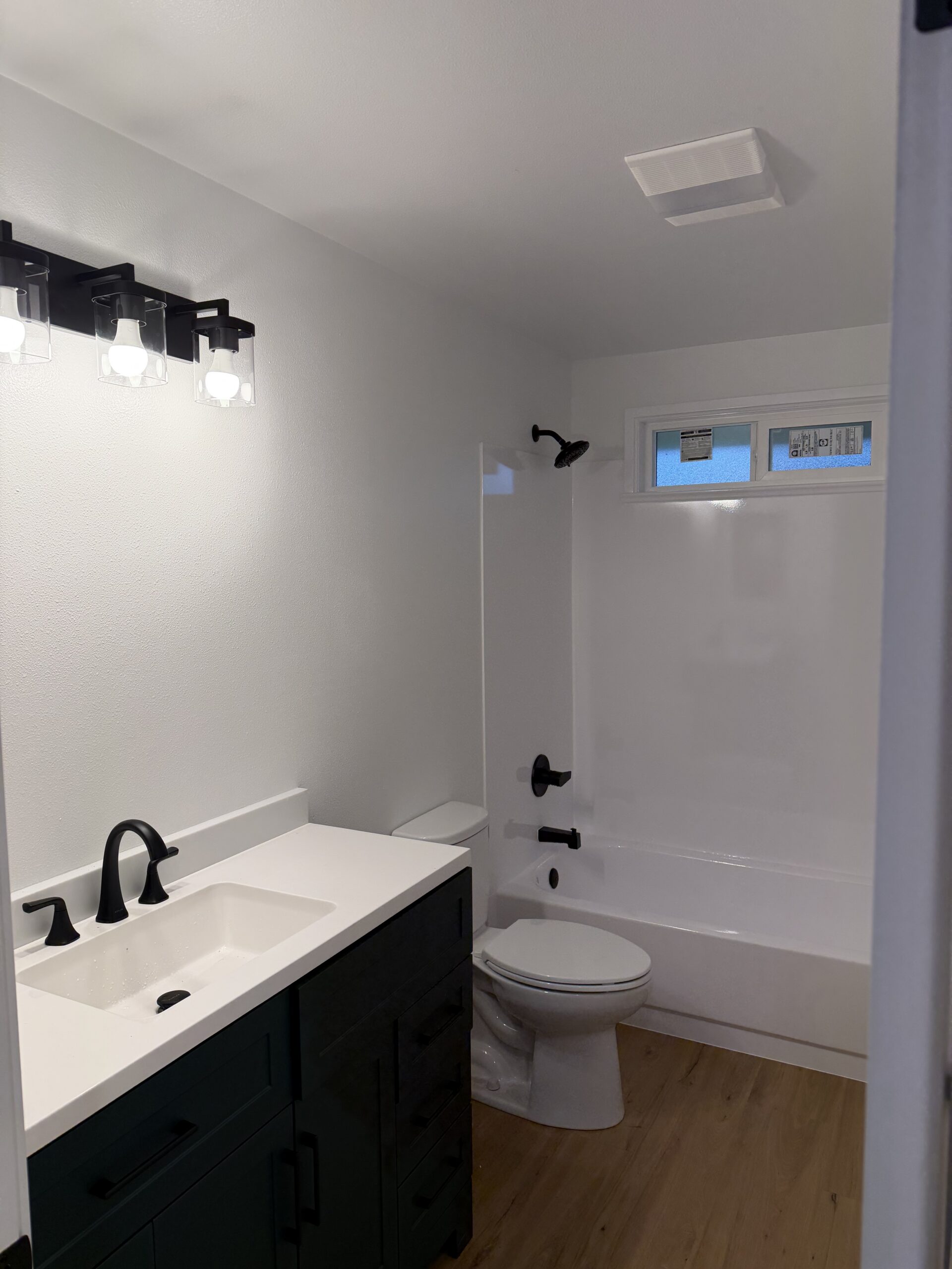 Modern bathroom with a white sink, black fixtures, dark vanity, toilet, and a white bathtub with shower. Wood-style flooring and overhead lighting are also visible.
