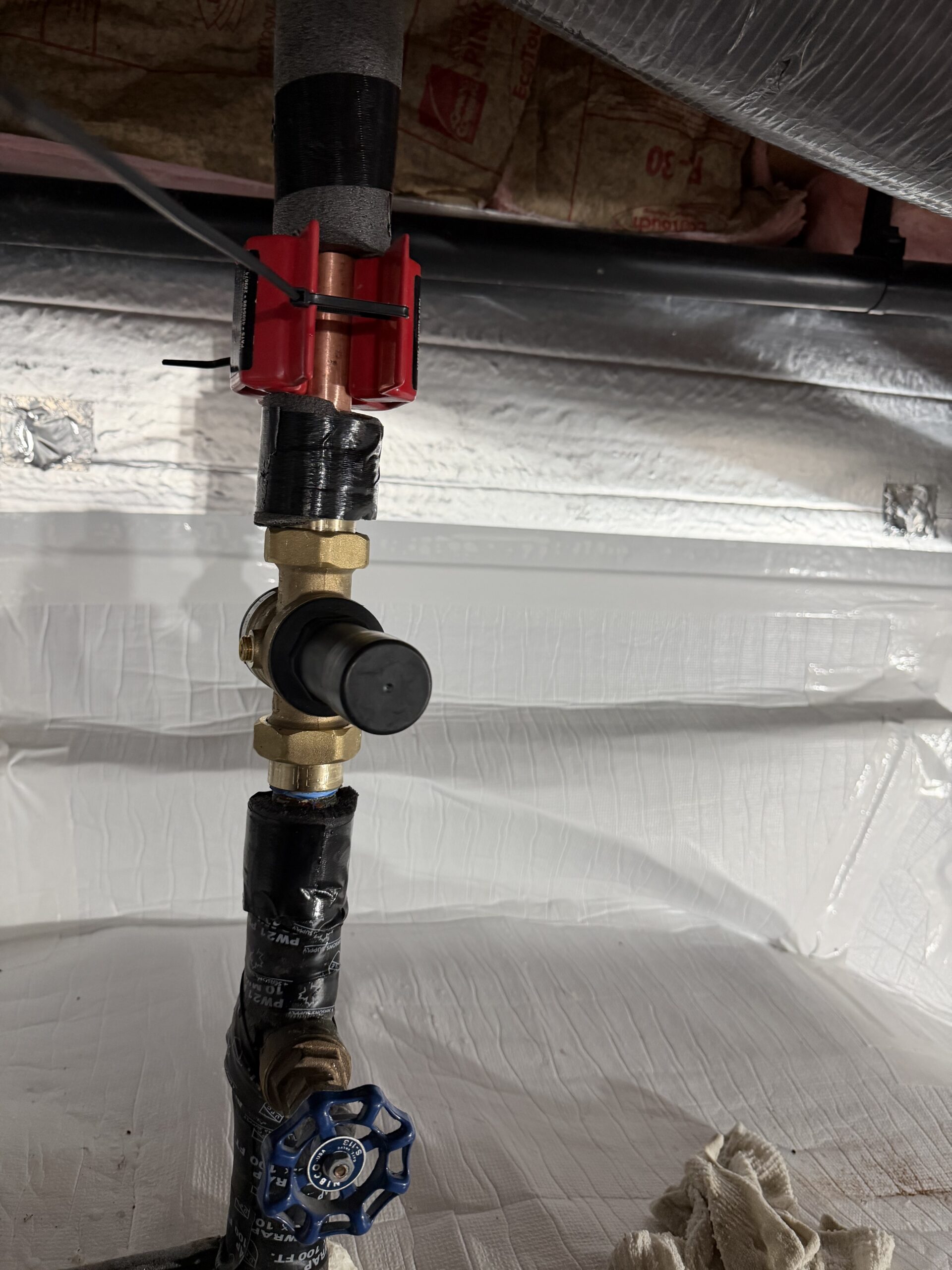 Close-up of insulated copper and black pipes with a brass valve assembly, black handle, and blue shut-off valve in a basement or crawl space.