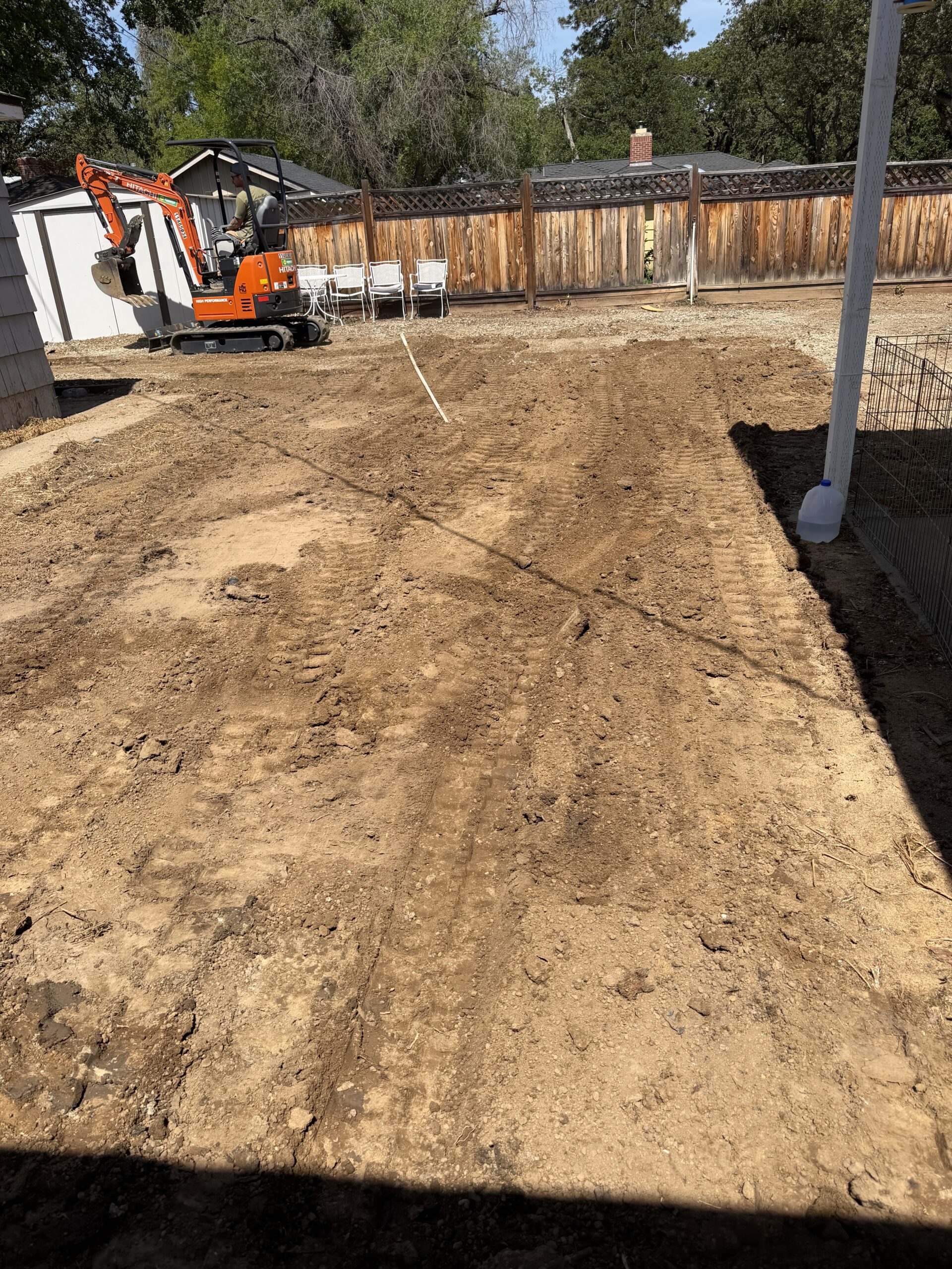 A dirt yard with visible tire tracks, a small orange excavator, a wooden fence in the background, and stacked white chairs near the fence.