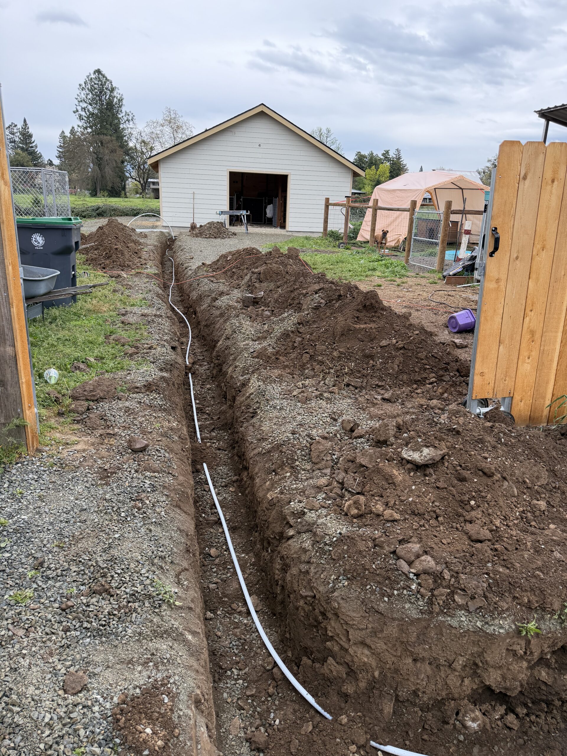 A long trench with a white pipe runs through a yard toward a detached garage, with dirt piles on each side and a wooden fence in the foreground.