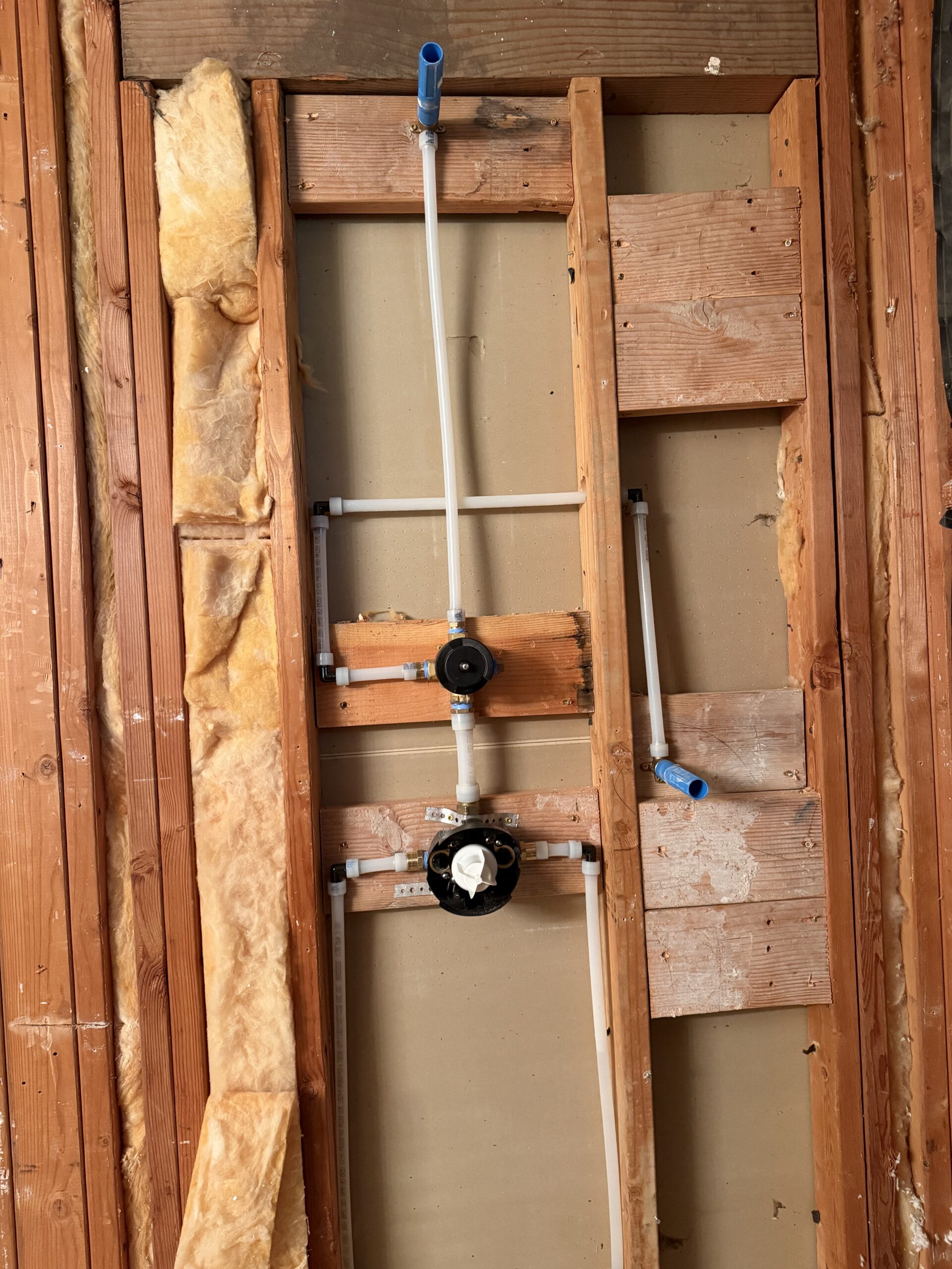Exposed wall with new plumbing installation, showing white pipes, valves, and unfinished wood framing during a bathroom renovation.