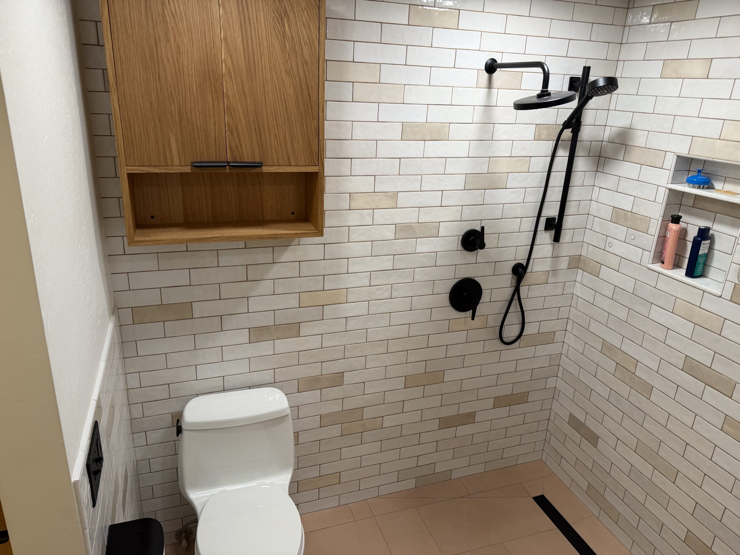 Bathroom with a white toilet, wooden cabinet, walk-in shower with black fixtures, and wall-mounted shelves holding toiletries against white subway tiles.